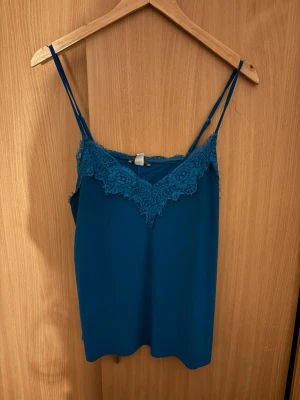 Blå spetslinne från Amisu - Snyggt blått linne från Amisu med tunna axelband och spetsdetaljer längs v-ringningen. Perfekt att styla till jeans eller kjol för en trendig look. Materialet känns mjukt och lätt, och spetsen ger en extra touch av stil.