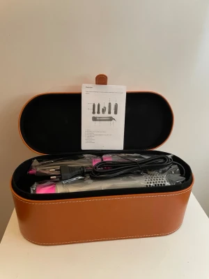 Multistyler hårverktyg med tillbehör - Snygg multistyler för hår med flera utbytbara tillbehör, perfekt för att skapa olika frisyrer. Kommer i en elegant brun förvaringsväska med vita sömmar. Verktyget har silver- och rosa detaljer och flera borsthuvuden i plast och metall.