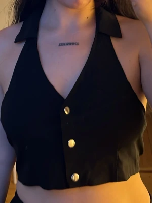 Svart croppad linnetopp med krage - Snygg svart croppad linnetopp med krage och guldknappar framtill. Toppen är ärmlös och har djup urringning samt öppen rygg, vilket ger en trendig och edgy look. Perfekt för dig som vill sticka ut lite extra.