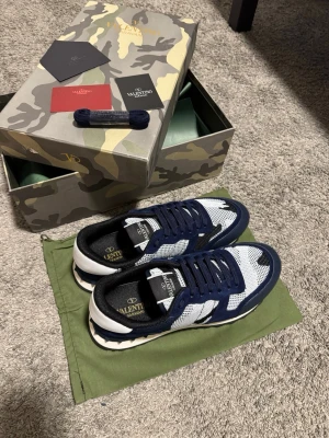 Valentino Garavani blåvita sneakers - Snygga sneakers från Valentino Garavani i blått, vitt och svart med camomönster. Ovandelen är i mocka och sulan har coola nitar baktill. Skorna kommer i originalkartong, dustbag och extra skosnören. Strl 44