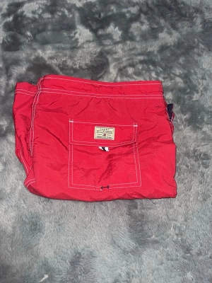 Tommy Hilfiger Badbyxor – Röda (Nyskick) - Säljer ett par fräscha röda badshorts från Tommy Hilfiger. Fina marina detaljer och vita sömmar. Oanvända och nytvättade med alla tvättlappar kvar.  • Skick: 10/10 – Nyskick, inga defekter. • Detaljer: Bakficka med tryckknapp och innerbyxa i nät. Kolla in mina andra annonser!