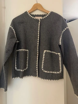 Grå kofta med vita detaljer från Nelly - Säljer en grå kofta från Nelly med vita, sicksackformade kanter längs halsringning, framkant, ärmar och fickor. Koftan har två stora fickor framtill och stängs med en knapp upptill. Perfekt för dig som gillar en unik och stilren look.