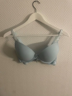 Ljusblå push-up bh med spetsdetaljer - Snygg ljusblå push-up bh med vadderade kupor och spetsdetaljer längs sidorna och bak. Justerbara axelband och liten rosett framtill. Perfekt för dig som vill ha en feminin och stilren look.