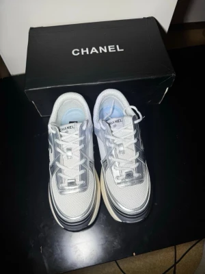 Chanel vita och silver sneakers - Snygga Chanel sneakers i vitt och silver med klassisk CC-logga på sidan. Ovandelen är i mesh och läder, med vita skosnören och en chunky sula i vitt och svart. Perfekta för dig som vill ha en exklusiv och trendig look.