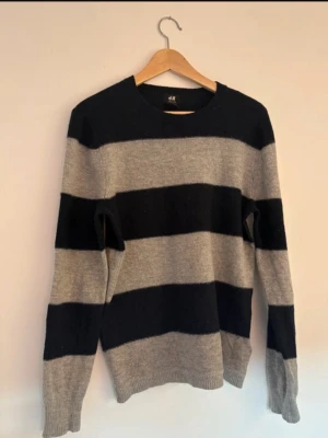 Randig stickad tröja  - Stickad tröja från H&M med breda horisontella ränder i svart och ljusgrått. Tröjan har rund halsringning och långa ärmar. Perfekt för dig som gillar en enkel men snygg stil. Materialet känns mjukt och bekvämt.