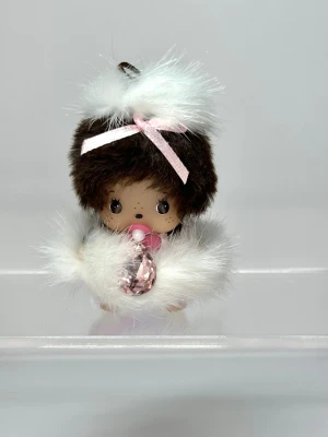 Söt Monchhichi figur med fluff - En gullig Monchhichi figur med brunt hår, vit fluffig mössa och jacka, samt en rosa rosett och napp. Figurer håller en stor rosa kristall och har detaljerade ögon och fräknar. Perfekt som samlarobjekt eller dekoration.