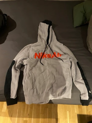 Grå Nike Air hoodie med svart detalj - Snygg grå hoodie från Nike Air med stor röd logga på bröstet. Svarta detaljer på ärmar och huva, samt dragsko i huvan. Perfekt för en sportig och avslappnad stil. Mjuk bomullsblandning och känguruficka framtill.