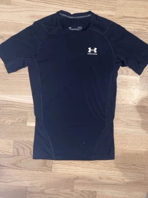 Svart kompressionströja Under Armour - Säljer en svart kompressionströja från Under Armour med HeatGear-teknologi. Tröjan har korta ärmar, rund hals och vit logga på bröstet. Perfekt för träning och sportaktiviteter där du vill hålla dig sval och bekväm.