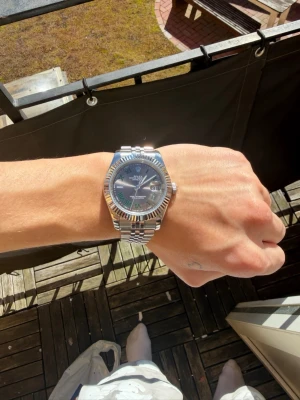  silver klocka - Snygg med silverfärgat armband i metall och grå urtavla. Klockan har romerska siffror i grönt och datumvisning. Klassisk räfflad bezel och elegant design som sticker ut på handleden.