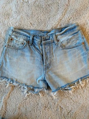Ljusblå jeansshorts från Levi's - Säljer ett par ljusblå jeansshorts från Levi's med fransig rå kant och klassisk femficksmodell. Shortsen har normal passform och är tillverkade i bomull. Perfekta för varma dagar och en avslappnad stil. Kolla alla bilder för defekter