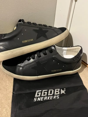 Svarta Golden Goose sneakers med glitter - Snygga svarta sneakers från Golden Goose med ikonisk stjärndetalj på sidan och glitter på hälen. Skorna har vita slitna sulor och GGDB/SSTAR-tryck i guld. Tillverkade i läder med svarta skosnören och vit insida. Perfekta för dig som gillar streetstyle och unika detaljer.