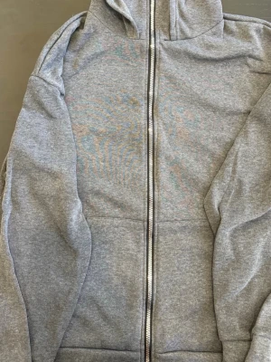 Grå zip hoodie med huva - En klassisk grå zip hoodie med dragkedja framtill och stor huva. Hoodien har två fickor på magen och är gjord i mjukt sweatshirtmaterial, perfekt för en avslappnad stil. Passar till det mesta och är superbekväm.