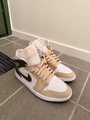 Air Jordan 1 Mid beige/olivgrön - Snygga Air Jordan 1 Mid sneakers i beige, olivgrönt och vitt. Skorna har klassisk Jordan-logga på sidan, perforerad tåbox och beige snörning. Tillverkade i en mix av läder och syntetmaterial för en clean och modern look.