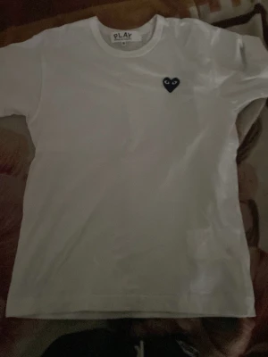 Vit Comme des Garçons Play t-shirt - Säljer en klassisk vit t-shirt från Comme des Garçons Play med det ikoniska svarta hjärtat broderat på bröstet. T-shirten är kortärmad och har en rund halsringning. Tillverkad i mjuk bomull för skön passform.