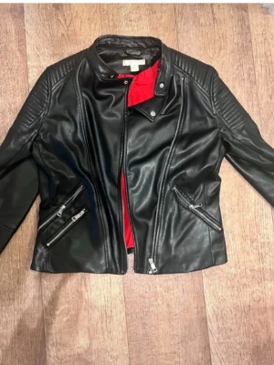 Svart skinnjacka - Snygg svart skinnjacka i bikerstil med coola detaljer som quiltade axelpartier och två snedställda dragkedjefickor. Jackan har en ståkrage med tryckknapp och röd innerfoder som ger en extra edge. Perfekt för dig som vill ha en stilren men tuff look.