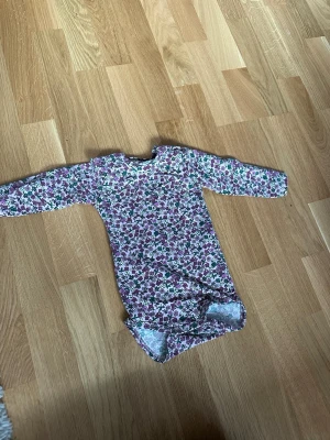 Blommig långärmad body - Söt långärmad body med blommigt mönster i lila, grönt och vitt. Rund halsringning och tryckknappar i grenen för enkel påklädning. Perfekt för dig som gillar färgglada och lekfulla plagg.