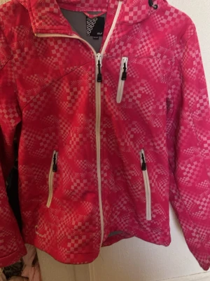 Rosa vindjacka från Stuf med mönster - Snygg rosa vindjacka från Stuf med schackrutigt mönster i olika nyanser av rosa och vitt. Jackan har vit dragkedja framtill, tre fickor med dragkedja och huva. Perfekt för blåsiga dagar och enkel att matcha med jeans eller träningskläder.