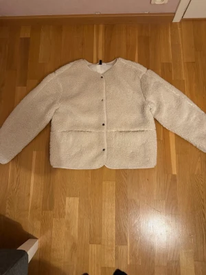 Beige teddyjacka från H&M - Mysig beige teddyjacka från H&M med rund halsringning och tryckknappar framtill. Jackan har två stora fickor och är gjord i fluffigt teddy-material som ger en chill och trendig look.