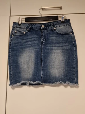 Blå jeanskjol med fransig kant - Snygg blå jeanskjol i kort modell med slitna detaljer och fransig nederkant. Kjolen har klassiska fem fickor, bälteshällor och knappstängning fram. Perfekt för en avslappnad och trendig look.