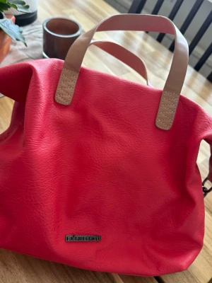 Rosa handväska från Björn Borg - Snygg rosa handväska från Björn Borg i konstläder med ljusbeige handtag. Väskan har en rymlig insida med dragkedja och innerfack, perfekt för att bära med sig det viktigaste. Stilsäker och enkel att matcha till olika outfits.