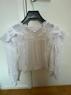 Vit volangblus från Gina Tricot - Söt vit blus från Gina Tricot med volangdetaljer och v-ringning. Blusen har långa ärmar och är gjord i ett tunt, luftigt tyg med struktur.