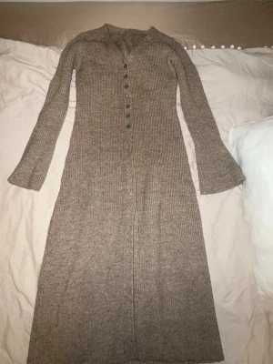 Lång ribbstickad kofta med knappar - Säljer en lång, ribbstickad kofta i beige/brun nyans med v-ringning och knappar hela vägen framtill. Koftan har långa ärmar med lätt utsvängda ärmslut och är gjord i ett mjukt, stickat material. Perfekt att slänga över jeans för en chill look.