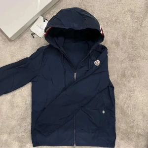 Marinblå vindjacka från Moncler - Snygg marinblå vindjacka från Moncler med huva och dragkedja framtill. Jackan har en liten Moncler-logga på ärmen och en rödvit rand på huvan. Perfekt för blåsiga dagar och enkel att matcha med det mesta.