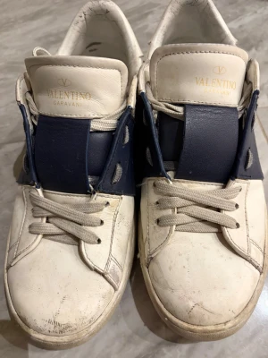 Valentino Garavani vita sneakers - Snygga vita sneakers från Valentino Garavani med breda mörkblåa detaljer över vristen. Skorna har klassisk låg modell, vita snören och är tillverkade i läder. Perfekta för dig som gillar exklusiva och trendiga märken.