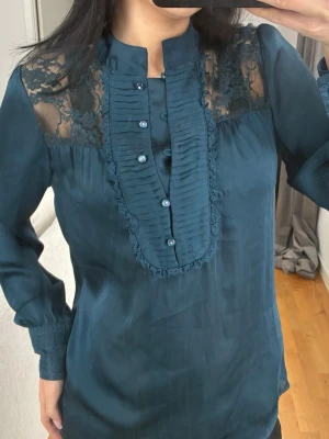 Mörkblå blus med spetsdetaljer - Snygg mörkblå blus med lång ärm och dekorativa spetsdetaljer över axlarna. Blusen har ståkrage, knappar framtill och plisserad panel med spetskant. Materialet är glansigt och ger en elegant känsla. Perfekt för dig som vill ha något extra till din outfit.
