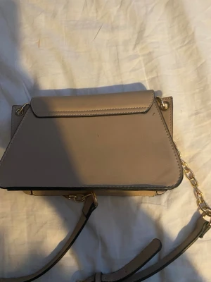 Beige axelväska med guldkedja - Snygg beige axelväska i konstläder med guldfärgade kedjedetaljer och justerbar axelrem. Väskan har en unik geometrisk form och ett dekorativt spänne framtill. Perfekt för dig som vill ha en stilren och trendig accessoar.