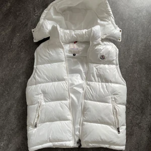 Vit väst från Moncler - Snygg vit väst från Moncler med avtagbar huva och två dragkedjefickor framtill. Västen har klassisk Moncler-logga på bröstet och quiltad design. Perfekt för lager-på-lager och håller dig varm under kyliga dagar.