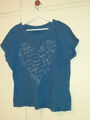 Blå t-shirt med texttryck frän Next - Blå t-shirt med korta ärmar och rund halsringning. Framsidan har ett vitt handskrivet texttryck i hjärtform. T-shirten är i mjuk bomull och har en avslappnad passform, perfekt för en chill och casual stil.