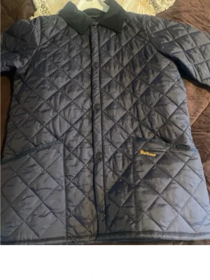  vindjacka Barbour - Snygg marinblå vindjacka från Barbour med quiltat mönster, tryckknappar framtill och två stora framfickor. Klassisk krage i kontrasttyg och diskret Barbour-logga på fickan. Perfekt för dig som gillar stilren och tidlös design.