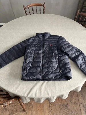 Svart pufferjacka från Ralph Lauren - Snygg svart pufferjacka från Ralph Lauren med diskret röd logga på bröstet. Jackan har dragkedja framtill, två sidofickor med dragkedja och en hög krage. Perfekt för kalla vinterdagar och ger en stilren look. Funkar lika bra på höst och vår.