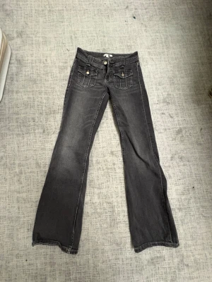 Grå bootcut jeans med fickdetaljer - Säljer ett par grå bootcut jeans med dubbla knappar och fickor framtill med lock och knapp. Jeansen har klassiska bakfickor och markerade sömmar. Materialet är denim med lätt tvättad look. Perfekta för dig som gillar en avslappnad men ändå trendig stil.