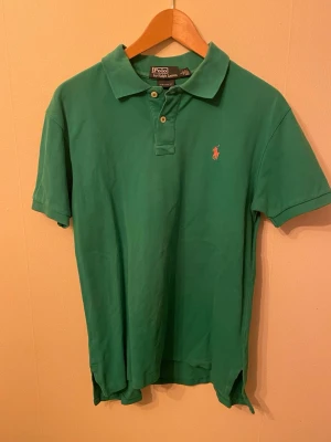 Grön pikétröja från Ralph Lauren - Snygg grön pikétröja från Polo Ralph Lauren med klassisk krage och två knappar framtill. Tröjan har den ikoniska broderade loggan i rött på bröstet och är tillverkad i bomull för en bekväm känsla. Perfekt till jeans eller shorts för en clean look.
