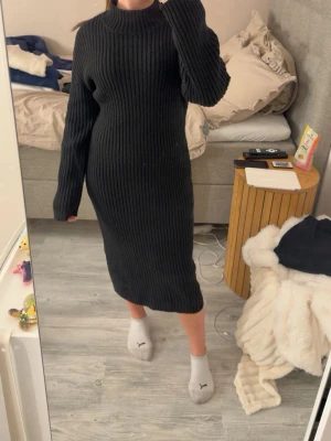 Svart ribbad stickad långklänning - Mysig svart stickad långklänning med ribbad struktur och lång ärm. Klänningen har en hög hals och rak passform som ger en clean och stilren look. Perfekt för dig som gillar enkel men trendig stil.