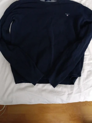 Mörkblå tröja från GANT - Snygg mörkblå långärmad tröja från GANT med diskret logga på bröstet. Tröjan har rund halsringning och är tillverkad i ett mjukt, tunt stickat material som känns skönt mot huden. Perfekt för dig som gillar klassisk och stilren design.