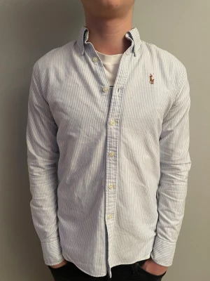 Ralph Lauren Skjorta - Vit och ljusblå randig skjorta från Ralph Lauren med klassisk krage och knappar framtill. Långärmad modell med broderad logga på bröstet. Jag är 170 lång och väger 57 kg, den passar perfekt på mig! Skjortan är i bomull och har en normal passform, perfekt för en clean och stilren look.