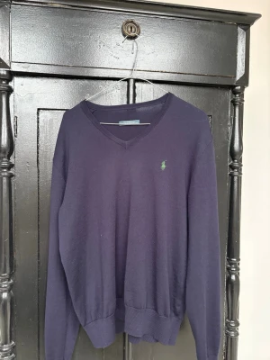 Marinblå stickad tröja från Ralph Lauren - Snygg marinblå stickad tröja från Ralph Lauren med klassisk v-ringning och den ikoniska gröna polospelaren broderad på bröstet. Tröjan har långa ärmar och ribbade muddar vid ärmslut och nederkant. Perfekt för lager på lager och stilren look.