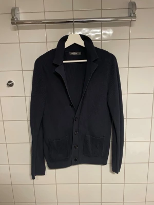 Mörkblå stickad kofta med fickor - Snygg mörkblå stickad kofta från Dressmann med knappar framtill och två stora fickor. Koftan har en klassisk krage och är ribbstickad för en stilren look. Perfekt att slänga på sig över en t-shirt eller skjorta.