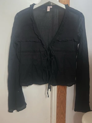 Svart blus med snörning och volang - Svart blus med v-ringning, snörning framtill och volangdetaljer längs halsen. Långärmad och tillverkad i ett tunt, lätt material som ger en luftig känsla. Perfekt för dig som gillar en romantisk och trendig look.💗 Aldrig använd!!