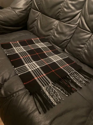 Svart rutig halsduk med fransar - Snygg svart halsduk med klassiskt rutmönster i vitt och tunna röda linjer. Halsduken har långa fransar i ändarna och är tillverkad i ett mjukt tyg som känns skönt mot huden. Perfekt accessoar för att lyfta din outfit under kyliga dagar.