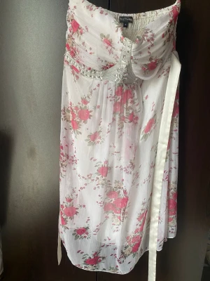 Vit blommig sommarklänning med små stenar  - Med rosa blommigt mönster och dekorativa stenar framtill. Klänningen har smock upptill för skön passform och ett tunt, luftigt tyg som ger en romantisk känsla. Perfekt för varma dagar och somriga tillfällen.