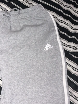 Gråa mjukisbyxor från Adidas - Säljer ett par gråa mjukisbyxor från Adidas med vita ränder längs sidorna och dragsko i midjan. Byxorna har en avslappnad passform och är tillverkade i mjukt material, perfekta för chill dagar. Adidas-logga på vänster ben. Det står att det är L men det stämmer inte utan det är som en lite kortare S