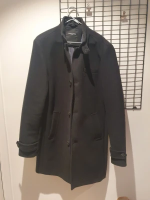 Svart ullrock från Jack & Jones - Knappt använd. Nypris 1800:-.               Stilren svart rock från Jack & Jones i ullblandning. Klassisk rak modell med knäppning framtill, ståkrage med knappdetalj och två sidofickor.