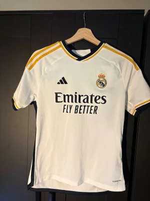 Real Madrid matchtröja Adidas - Säljer en vit Real Madrid matchtröja från Adidas med klubbens emblem på bröstet och sponsorlogga 'Emirates Fly Better'. Tröjan har gula och svarta detaljer på axlar och ärmslut, samt texten '¡Hala Madrid!' i nacken. Materialet är lätt och andas, perfekt för fotboll.