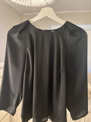 Svart satinblus med trekvartsärm - Stilren svart blus från NA-KD i glansigt satinmaterial. Blusen har rund halsringning, puffiga axlar och trekvartsärmar som ger en trendig siluett. Perfekt att matcha med både jeans och kjol för en snygg look.