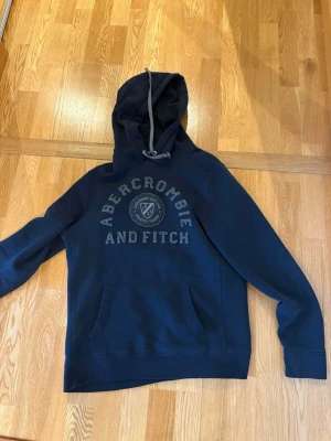 Mörkblå hoodie Abercrombie & Fitch - Snygg mörkblå hoodie från Abercrombie & Fitch med stor logotyp på bröstet och klassisk huva med grå dragsko. Hoodien har en stor magficka och är tillverkad i mjukt material som känns riktigt skönt. Perfekt för dig som gillar en avslappnad och cool stil.