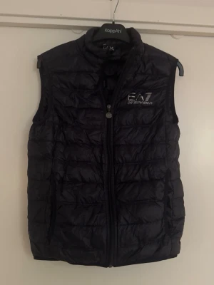 Emporio Armani EA7 väst - Snygg svart dunväst från Emporio Armani EA7 med quiltad design och dragkedja framtill. Västen har en hög krage och logga broderad på bröstet. Perfekt för lager på lager och ger en sportig vibe till din outfit.